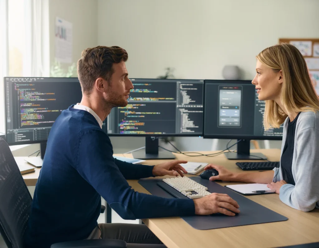 Lokale KI im Unternehmen: Was braucht man mindestens? 1 Zwei Personen besprechen Software an einem Arbeitsplatz mit mehreren Monitoren.